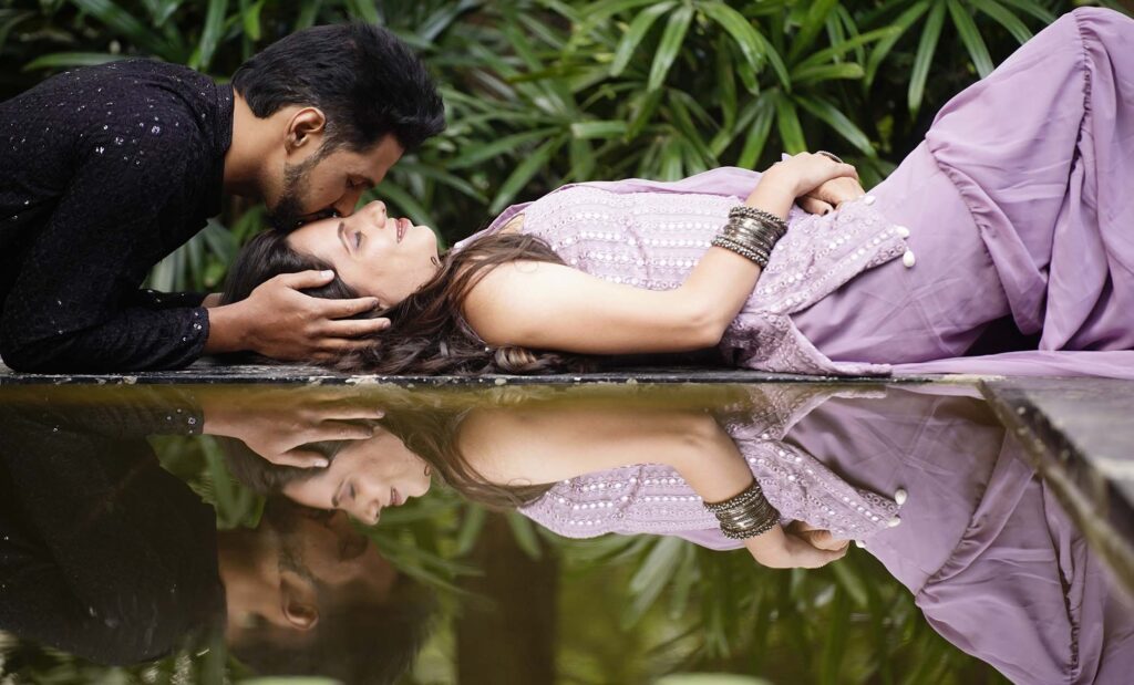 A romantic pre-wedding photo of a couple lying by a reflective water surface, surrounded by lush greenery. The man kissing woman's forehead, with their reflection beautifully mirrored in the water